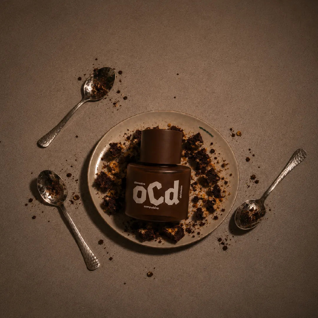 A brown bottle labeled 'oCd' sits on a beige plate surrounded by crumbs, with three silver spoons scattered on a textured brown surface.